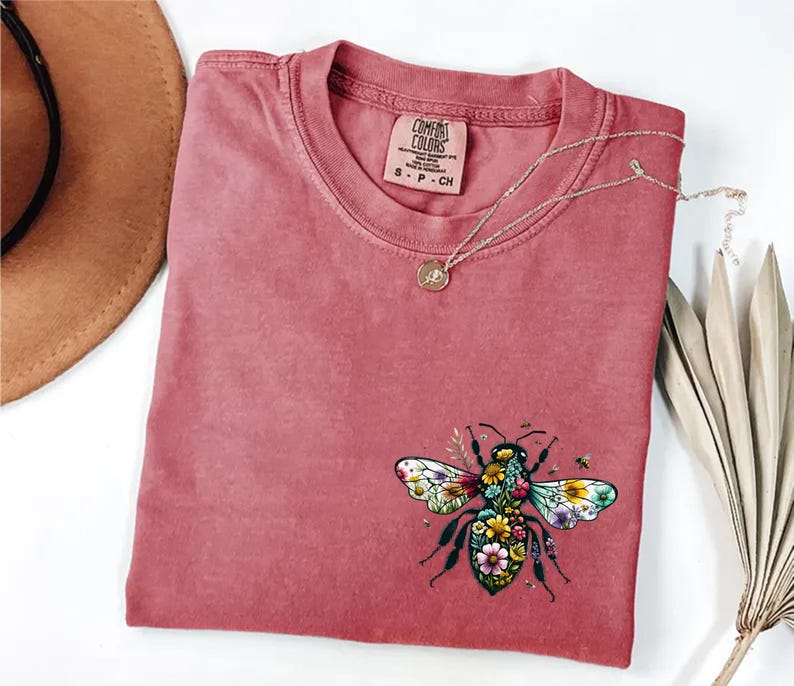May include: A dusty rose Comfort Colors t-shirt with a floral bee graphic. The bee design features colorful flowers and leaves. A gold necklace with a small pendant rests on the shirt. The shirt has a small Comfort Colors tag.