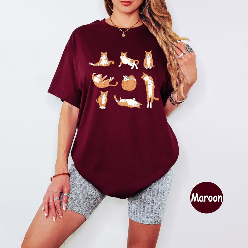 May include: Maroon t-shirt featuring a playful design of orange and white cats in various poses. The cats are arranged in a grid pattern. The shirt has short sleeves and a round neckline. The word "Maroon" is printed on the bottom right.