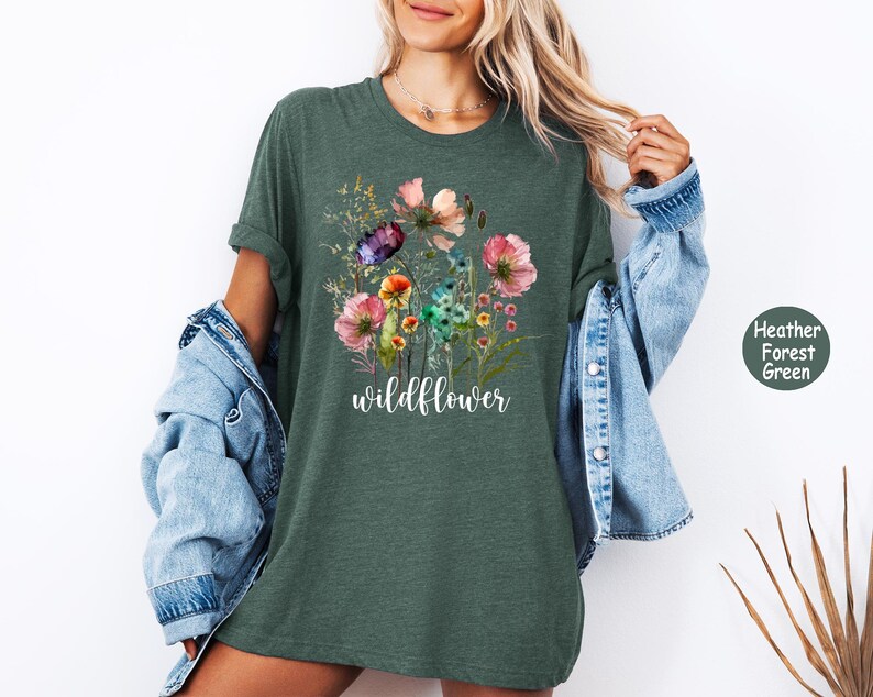 May include: Heather forest green t-shirt featuring a watercolor floral design with pink, purple, and yellow flowers. The word "wildflower" is written in a cursive font below the flowers. The shirt is a relaxed fit.