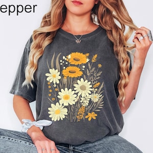 K&ouml;nnte beinhalten: Dunkelgraues T-Shirt mit einem Blumendesign mit gelben Sonnenblumen und wei&szlig;en G&auml;nsebl&uuml;mchen. Das Wort "Pepper" ist in der oberen linken Ecke aufgedruckt. Das Shirt hat eine lockere Passform.