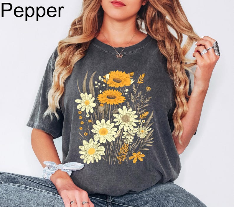 K&ouml;nnte beinhalten: Dunkelgraues T-Shirt mit einem Blumendesign mit gelben Sonnenblumen und wei&szlig;en G&auml;nsebl&uuml;mchen. Das Wort "Pepper" ist in der oberen linken Ecke aufgedruckt. Das Shirt hat eine lockere Passform.