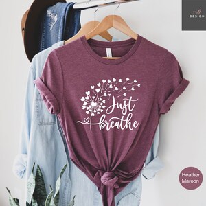 以下が含まれることがあります： ヘザーマルーンの半袖Tシャツ。ハート型の種が風に舞うタンポポの白いグラフィックが特徴です。タンポポのデザインの横には、白いスクリプトフォントで「Just breathe」という文字がプリントされています。