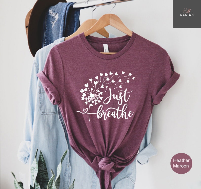 以下が含まれることがあります： ヘザーマルーンの半袖Tシャツ。ハート型の種が風に舞うタンポポの白いグラフィックが特徴です。タンポポのデザインの横には、白いスクリプトフォントで「Just breathe」という文字がプリントされています。