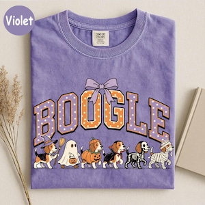 K&ouml;nnte beinhalten: Ein violettes Kurzarm-T-Shirt mit dem Schriftzug BOOGLE in orangefarbenen, wei&szlig; gepunkteten Buchstaben und einer violetten Schleife dar&uuml;ber. Unter dem Text sind sechs Beagle-Hunde in Halloween-Kost&uuml;men abgebildet: Hexe, Geist, K&uuml;rbis, Vampir, Skelett und Mumie.