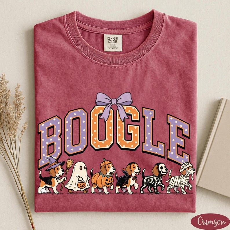 K&ouml;nnte beinhalten: Ein karmesinrotes T-Shirt mit Rundhalsausschnitt und dem Schriftzug BOOGLE in gro&szlig;en, gepunkteten College-Buchstaben, die von einer hellvioletten Schleife gekr&ouml;nt werden. Darunter befinden sich sechs Beagle-Hunde in Halloween-Kost&uuml;men: Hexe, Geist, K&uuml;rbis, Vampir, Skelett und Mumie.