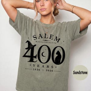 Puede incluir: Una camiseta de manga corta color verde salvia con un estampado gr&aacute;fico negro que dice Salem Massachusetts 400 Years 1626-2026. El dise&ntilde;o incluye un sombrero de bruja, una luna creciente dentro del cero y la silueta de un gato negro.
