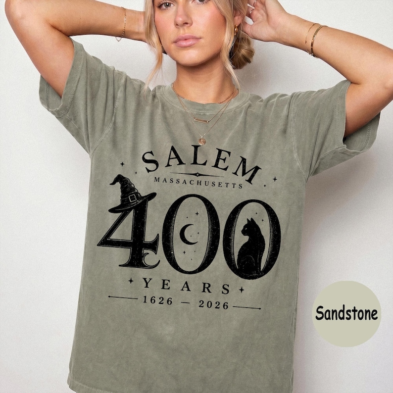 Puede incluir: Una camiseta de manga corta color verde salvia con un estampado gr&aacute;fico negro que dice Salem Massachusetts 400 Years 1626-2026. El dise&ntilde;o incluye un sombrero de bruja, una luna creciente dentro del cero y la silueta de un gato negro.