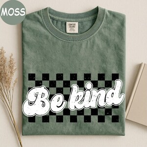 Pu&ograve; includere: Una t-shirt a maniche corte color verde muschio con un motivo a scacchi nero e verde al centro. Il testo "Be kind" &egrave; stampato in un carattere bianco in stile retr&ograve; con effetto usurato sopra il disegno a scacchi. Girocollo classico.