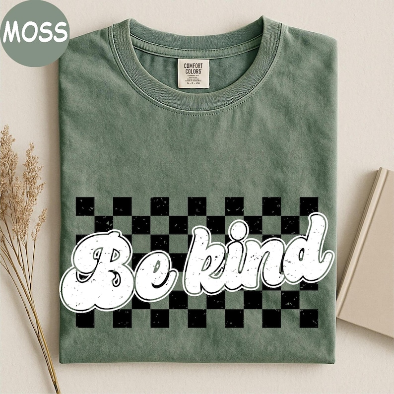 Pu&ograve; includere: Una t-shirt a maniche corte color verde muschio con un motivo a scacchi nero e verde al centro. Il testo "Be kind" &egrave; stampato in un carattere bianco in stile retr&ograve; con effetto usurato sopra il disegno a scacchi. Girocollo classico.