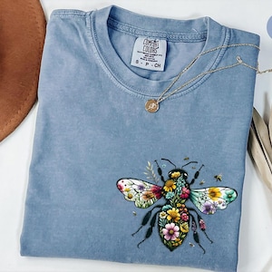 May include: Blue jean colored t-shirt with a floral bee graphic. The bee's wings are multi-colored, and the body is filled with flowers. A gold necklace with a pendant rests on the shirt. The shirt has a Comfort Colors tag.