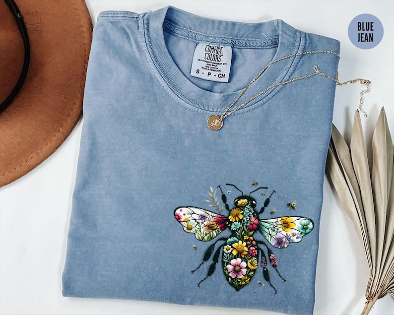 May include: Blue jean colored t-shirt with a floral bee graphic. The bee's wings are multi-colored, and the body is filled with flowers. A gold necklace with a pendant rests on the shirt. The shirt has a Comfort Colors tag.