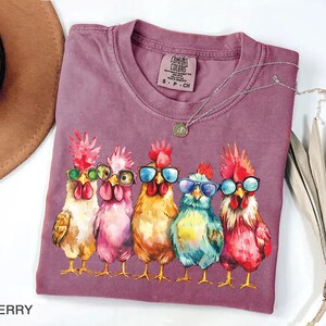 Puede incluir: Una camiseta morada con un gr&aacute;fico de cinco pollos con gafas de sol. Los pollos est&aacute;n dibujados en un estilo de dibujos animados con plumas y picos coloridos. El texto "Comfort Colors" est&aacute; impreso en la camiseta.