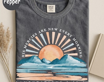 May include: A charcoal gray crew neck t-shirt featuring a graphic of a sun rising over mountains and water. The text reads His Mercies Are New Every Morning and Lamentations 3:22-23. The design uses muted tones of orange, blue, and cream.
