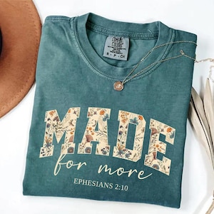 May include: Teal-colored t-shirt with the words "MADE for more" in a floral design. The text "Ephesians 2:10" is printed below. A gold necklace with a pendant is draped across the shirt.