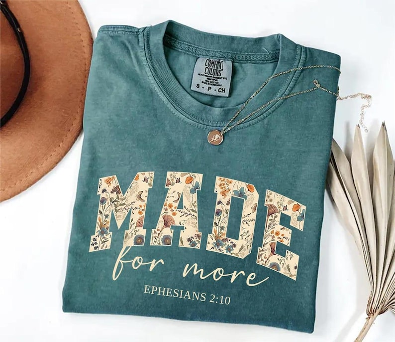 May include: Teal-colored t-shirt with the words "MADE for more" in a floral design. The text "Ephesians 2:10" is printed below. A gold necklace with a pendant is draped across the shirt.