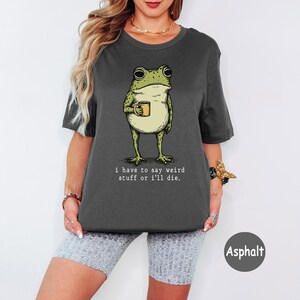 May include: A dark grey short-sleeve t-shirt featuring a graphic of a green frog holding a small yellow mug. Below the frog, the text reads, "i have to say weird stuff or i'll die." The shirt has a crew neckline and a relaxed, boxy fit.