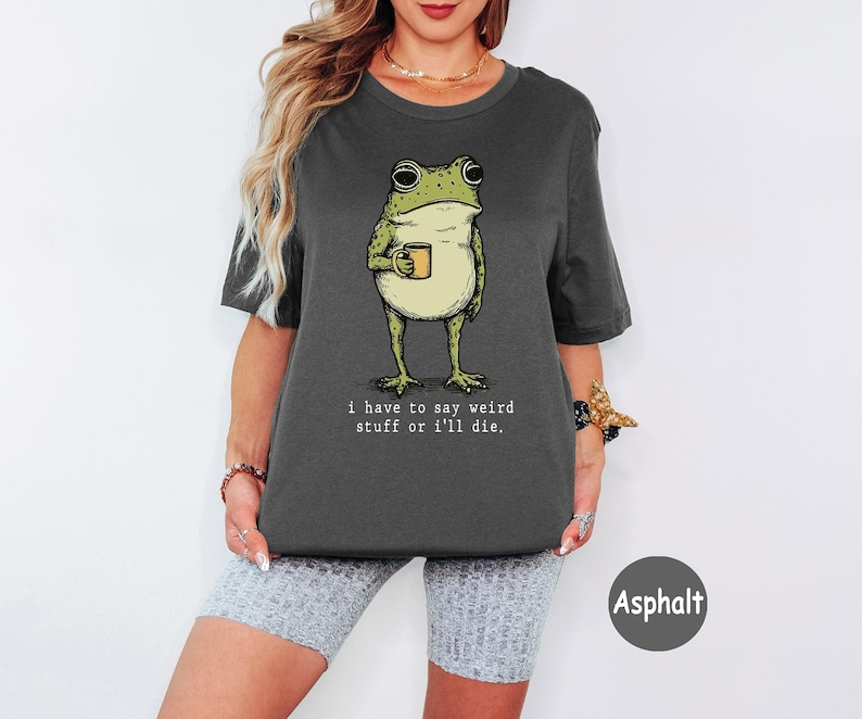 May include: A dark grey short-sleeve t-shirt featuring a graphic of a green frog holding a small yellow mug. Below the frog, the text reads, "i have to say weird stuff or i'll die." The shirt has a crew neckline and a relaxed, boxy fit.