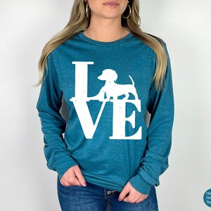 May include: Teal long-sleeve shirt with the word "LOVE" in white block letters, featuring a white dachshund dog silhouette. The shirt is made of a soft material and is suitable for casual wear.