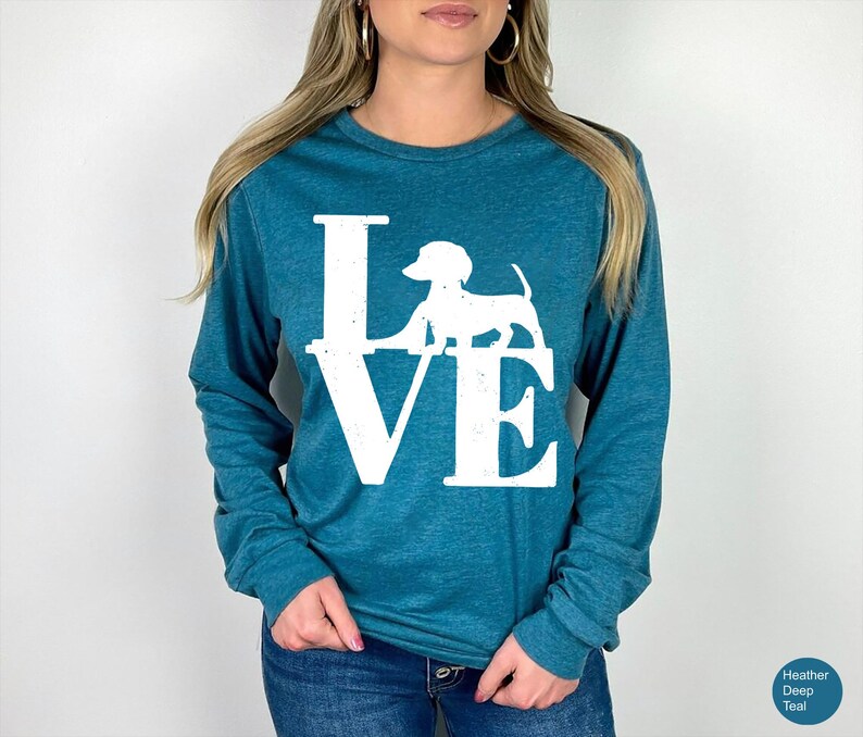 May include: Teal long-sleeve shirt with the word "LOVE" in white block letters, featuring a white dachshund dog silhouette. The shirt is made of a soft material and is suitable for casual wear.