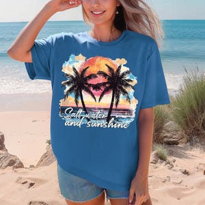 May include: A blue t-shirt featuring a sunset scene with palm trees and the text "Salt-water and sunshine." The graphic includes a vibrant orange and yellow sun, with the palm trees in silhouette.