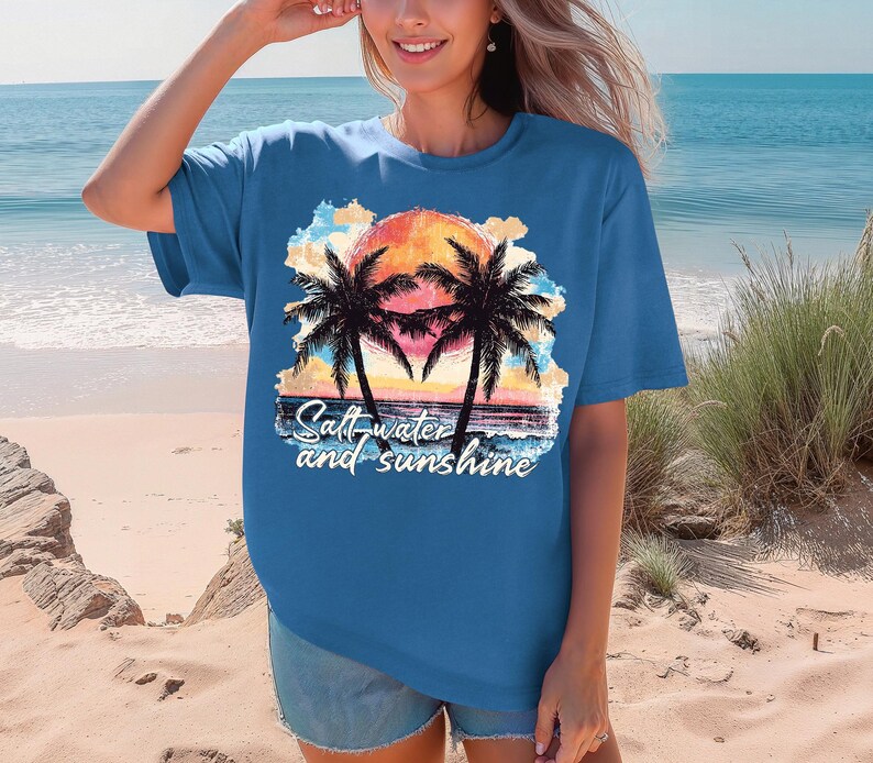May include: A blue t-shirt featuring a sunset scene with palm trees and the text "Salt-water and sunshine." The graphic includes a vibrant orange and yellow sun, with the palm trees in silhouette.