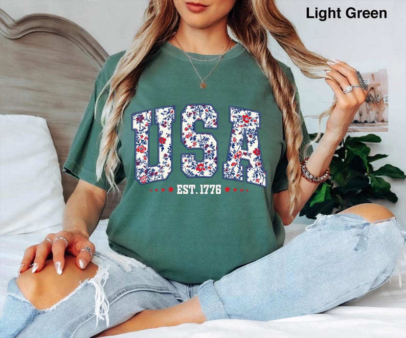 May include: Light green t-shirt with the word "USA" in a floral design, with "EST. 1776" below. The shirt is a relaxed fit and is worn with light blue jeans. The shirt is a light green color.