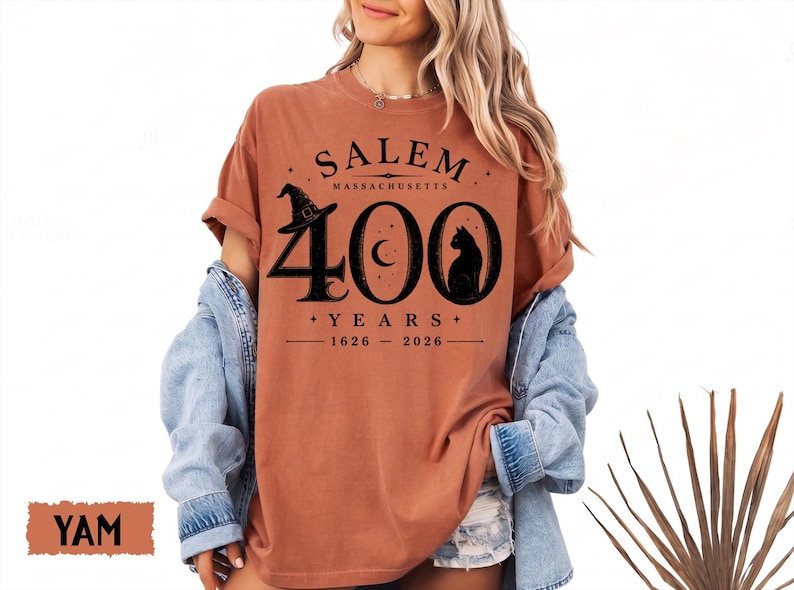 Puede incluir: Una camiseta de color &oacute;xido con un gr&aacute;fico negro que dice SALEM MASSACHUSETTS 400 YEARS 1626-2026. El dise&ntilde;o incluye un sombrero de bruja, una luna creciente y la silueta de un gato integrados en el n&uacute;mero 400, con la palabra YAM visible.