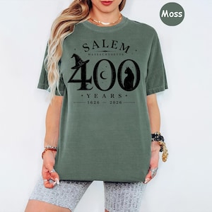 Puede incluir: Una camiseta de manga corta color verde musgo con un estampado gr&aacute;fico negro que dice SALEM MASSACHUSETTS 400 YEARS 1626-2026, acompa&ntilde;ado de ilustraciones de un sombrero de bruja, una luna creciente y la silueta de un gato en el pecho.