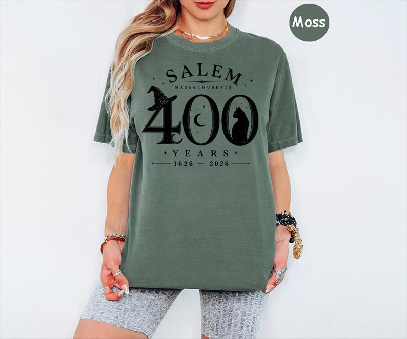 Puede incluir: Una camiseta de manga corta color verde musgo con un estampado gr&aacute;fico negro que dice SALEM MASSACHUSETTS 400 YEARS 1626-2026, acompa&ntilde;ado de ilustraciones de un sombrero de bruja, una luna creciente y la silueta de un gato en el pecho.