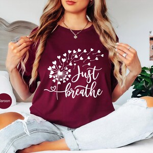 以下が含まれることがあります： マルーンカラーの半袖Tシャツ。ハート型の種が風に舞うタンポポの白いグラフィックが描かれています。シャツの前面には、タンポポのデザインの下に白い筆記体で「Just breathe」という文字がプリントされています。
