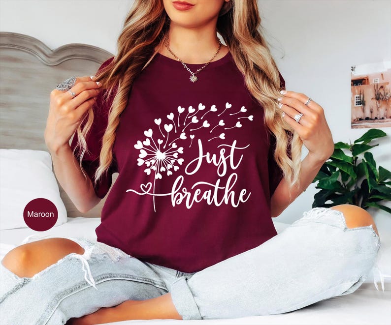 以下が含まれることがあります： マルーンカラーの半袖Tシャツ。ハート型の種が風に舞うタンポポの白いグラフィックが描かれています。シャツの前面には、タンポポのデザインの下に白い筆記体で「Just breathe」という文字がプリントされています。