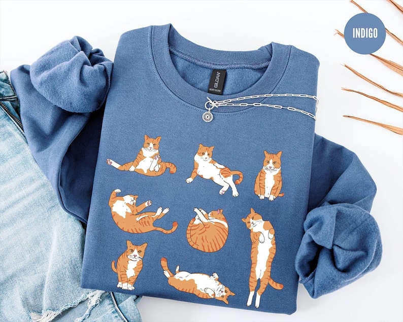 May include: An indigo blue crewneck sweatshirt featuring a graphic print of seven orange and white tabby cats in various lounging positions. The sweatshirt has a ribbed collar and long sleeves, with a small circular tag labeled INDIGO.