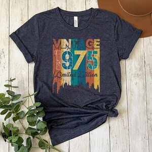May include: Heather navy blue t-shirt with a vintage design. The design features the word "VINTAGE" above the number "1975" and the words "Limited Edition" in a retro color scheme of orange, yellow, teal, and brown.