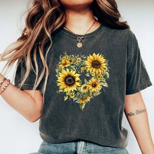 Pode incluir: T-shirt cinzenta escura com um design em forma de cora&ccedil;&atilde;o de girass&oacute;is amarelos. Os girass&oacute;is t&ecirc;m centros castanhos e folhas verdes. A t-shirt tem um aspeto vintage e desgastado.