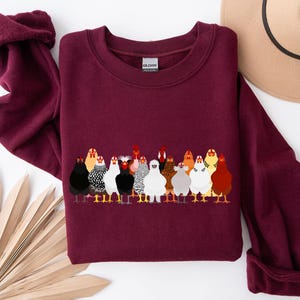 Peut inclure: Un sweat-shirt bordeaux avec une illustration color&eacute;e d'un groupe de poulets. Les poulets sont repr&eacute;sent&eacute;s dans diff&eacute;rentes couleurs et motifs, debout en rang. Un chapeau beige est visible en arri&egrave;re-plan.