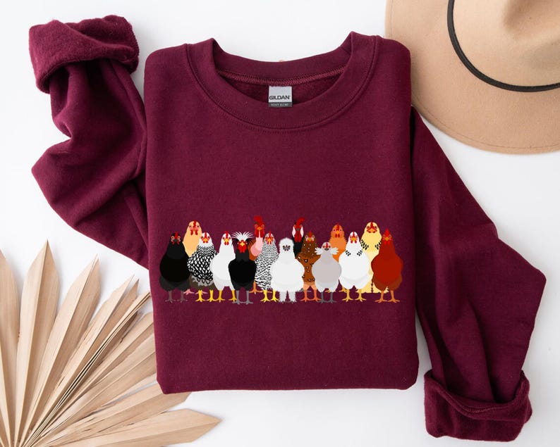 Peut inclure: Un sweat-shirt bordeaux avec une illustration color&eacute;e d'un groupe de poulets. Les poulets sont repr&eacute;sent&eacute;s dans diff&eacute;rentes couleurs et motifs, debout en rang. Un chapeau beige est visible en arri&egrave;re-plan.
