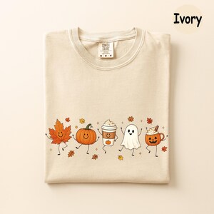 K&ouml;nnte beinhalten: Ein elfenbeinfarbenes T-Shirt mit einem Grafikdruck von einem l&auml;chelnden Ahornblatt, einem K&uuml;rbis, einem Kaffeebecher, einem Geist und einer Jack-o'-Lantern-Tasse mit Armen und Beinen. Das Shirt hat ein Comfort Colors-Etikett am Halsausschnitt.