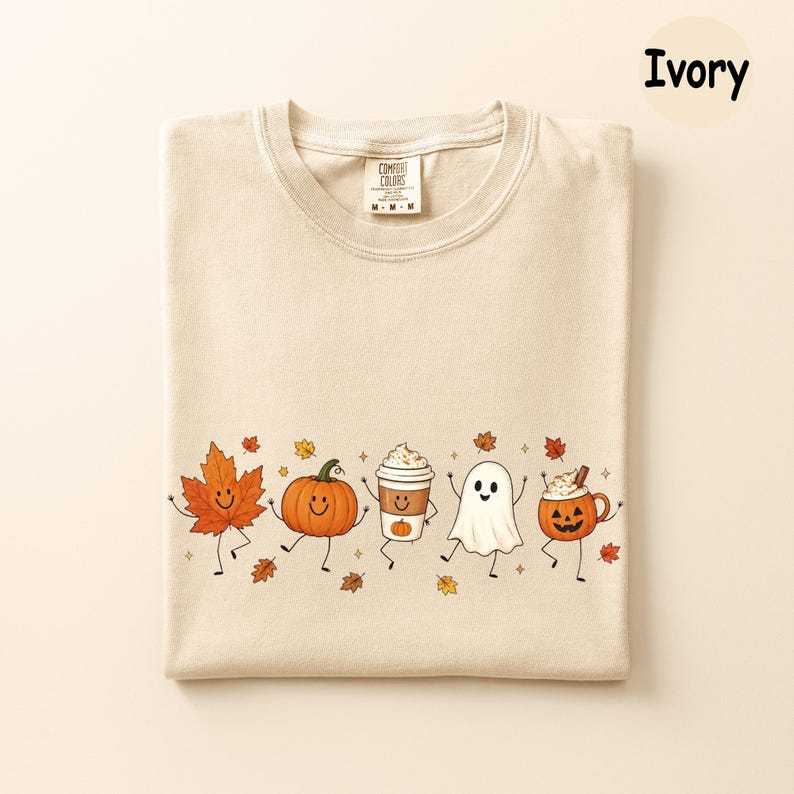 K&ouml;nnte beinhalten: Ein elfenbeinfarbenes T-Shirt mit einem Grafikdruck von einem l&auml;chelnden Ahornblatt, einem K&uuml;rbis, einem Kaffeebecher, einem Geist und einer Jack-o'-Lantern-Tasse mit Armen und Beinen. Das Shirt hat ein Comfort Colors-Etikett am Halsausschnitt.