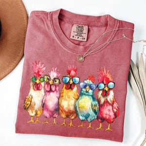Puede incluir: Una camiseta roja con un gr&aacute;fico de cinco pollos con gafas de sol. Los pollos est&aacute;n dibujados en un estilo de dibujos animados y llevan gafas de sol de diferentes colores. El texto "COMFORT COLORS" est&aacute; impreso en la camiseta.