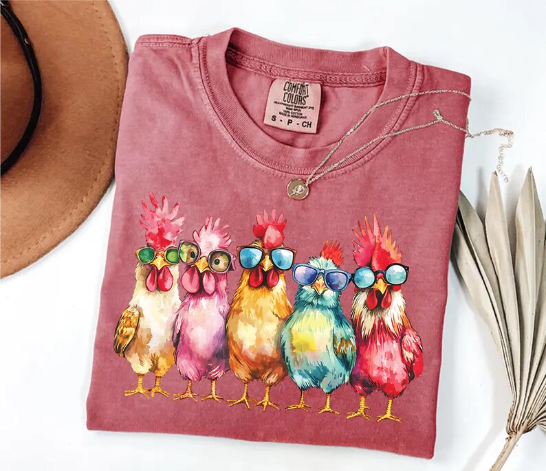 Puede incluir: Una camiseta roja con un gr&aacute;fico de cinco pollos con gafas de sol. Los pollos est&aacute;n dibujados en un estilo de dibujos animados y llevan gafas de sol de diferentes colores. El texto "COMFORT COLORS" est&aacute; impreso en la camiseta.