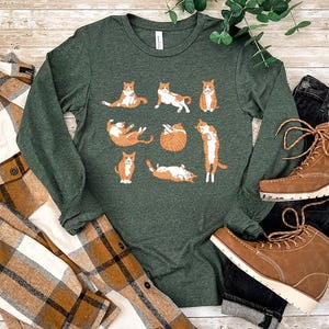 May include: A dark green long-sleeve crew neck shirt featuring a graphic print of eight orange tabby cats in various poses, including sitting, stretching, and curled up. The shirt is displayed alongside a brown plaid shirt and brown boots.