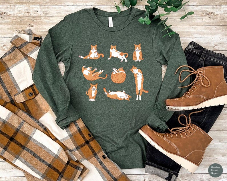 May include: A dark green long-sleeve crew neck shirt featuring a graphic print of eight orange tabby cats in various poses, including sitting, stretching, and curled up. The shirt is displayed alongside a brown plaid shirt and brown boots.