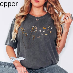 May include: A charcoal grey short-sleeve t-shirt featuring a graphic of three dandelion stems where the seeds are replaced by small, multi-coloured cats. The word Pepper is printed in the top left corner of the image in black text.