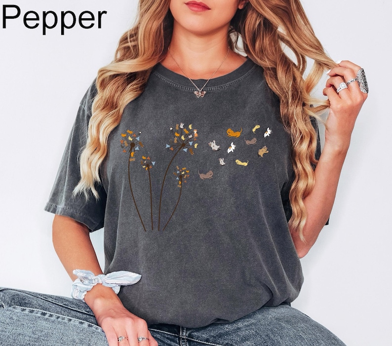 May include: A charcoal grey short-sleeve t-shirt featuring a graphic of three dandelion stems where the seeds are replaced by small, multi-coloured cats. The word Pepper is printed in the top left corner of the image in black text.