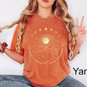 Pu&ograve; includere: Una maglietta arancione con un design celeste dorato raffigurante un sole, una luna e le fasi lunari. Il disegno &egrave; centrato sulla parte anteriore della maglietta. La parola "Yam" &egrave; stampata in nero in basso a destra.
