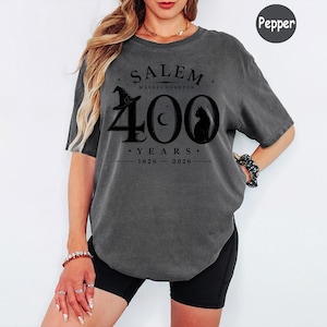 Puede incluir: Una camiseta de manga corta gris carb&oacute;n con un estampado gr&aacute;fico negro que dice SALEM MASSACHUSETTS 400 YEARS 1626-2026. El dise&ntilde;o incluye un sombrero de bruja, una luna creciente y la silueta de un gato integrada en el n&uacute;mero 400.