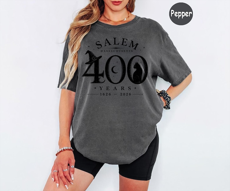 Puede incluir: Una camiseta de manga corta gris carb&oacute;n con un estampado gr&aacute;fico negro que dice SALEM MASSACHUSETTS 400 YEARS 1626-2026. El dise&ntilde;o incluye un sombrero de bruja, una luna creciente y la silueta de un gato integrada en el n&uacute;mero 400.