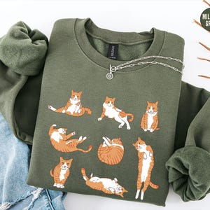 May include: A military green crewneck sweatshirt featuring a graphic print of seven orange and white tabby cats in various lounging positions. The sweatshirt has long sleeves and a ribbed collar, with a small circular tag reading Military Green.