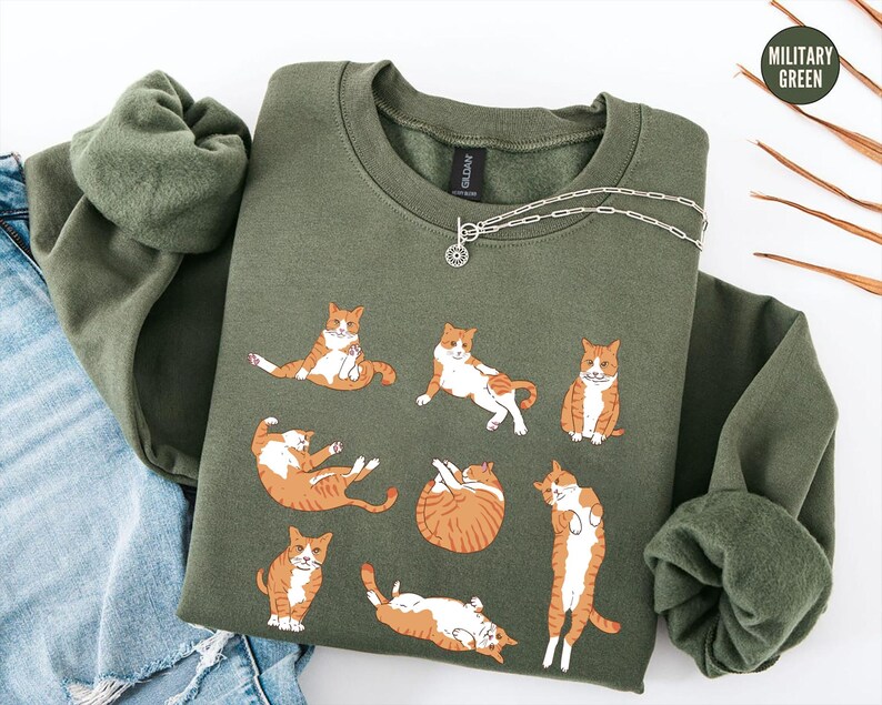 May include: A military green crewneck sweatshirt featuring a graphic print of seven orange and white tabby cats in various lounging positions. The sweatshirt has long sleeves and a ribbed collar, with a small circular tag reading Military Green.