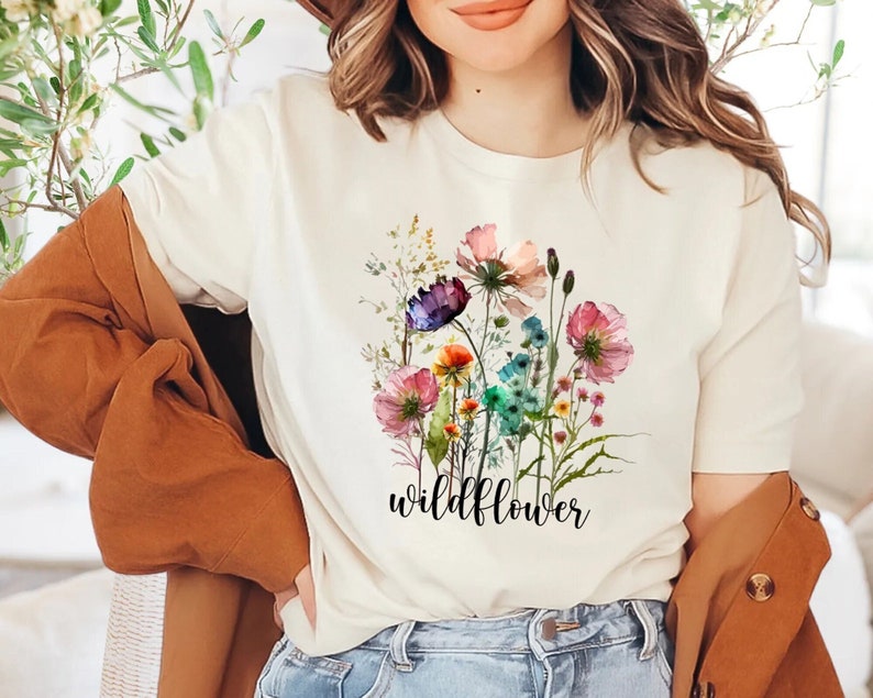 May include: Cream-colored t-shirt featuring a watercolor floral design with pink, purple, blue, and orange flowers. The word "wildflower" is printed in black script below the flowers. The shirt has short sleeves.
